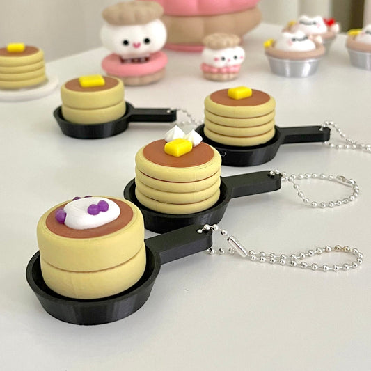 Miniature pancakes in a pan with syrup and a cherry on top, set against a neutral background.